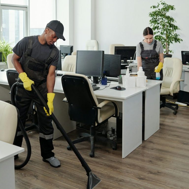 Two workers cleaning an office space, one using a vacuum and the other organizing supplies.